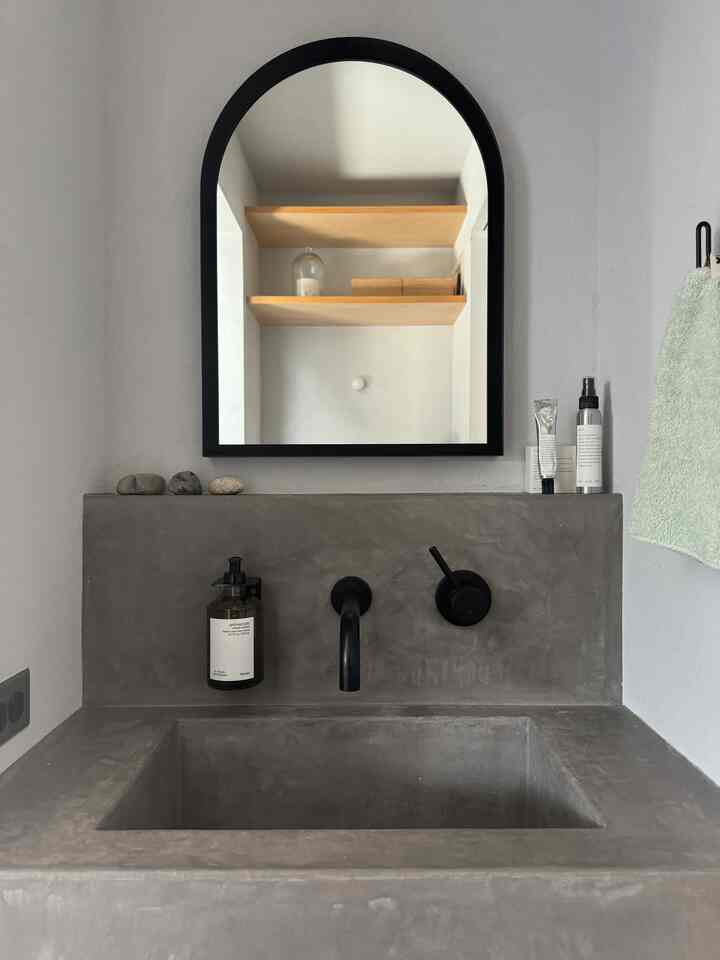 グレーのコンクリート製洗面台と黒いアーチ型鏡が配されたミニマルでシンプルなバスルームのインテリア