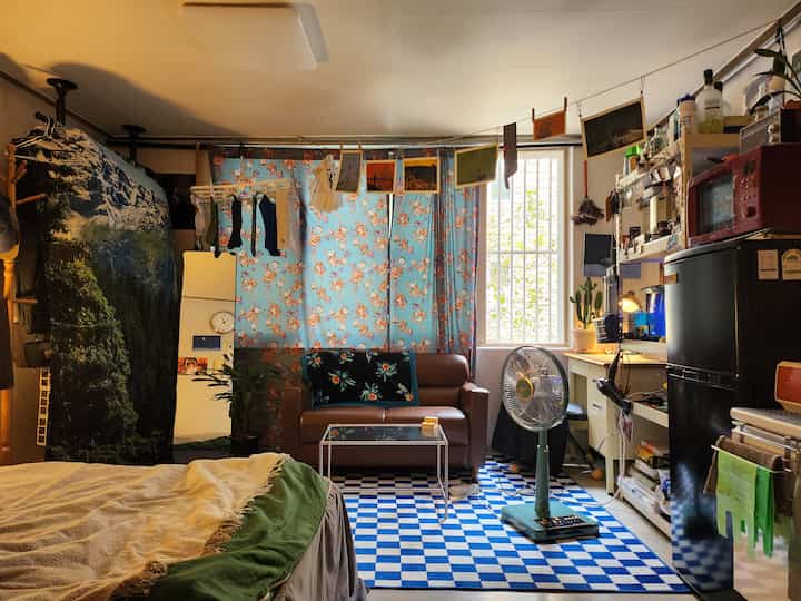 青い花柄のカーテンとチェック柄ラグが印象的な狭いワンルームのリビング兼寝室、茶色のレザーソファとヴィンテージ扇風機が配置された雰囲気のあるインテリア