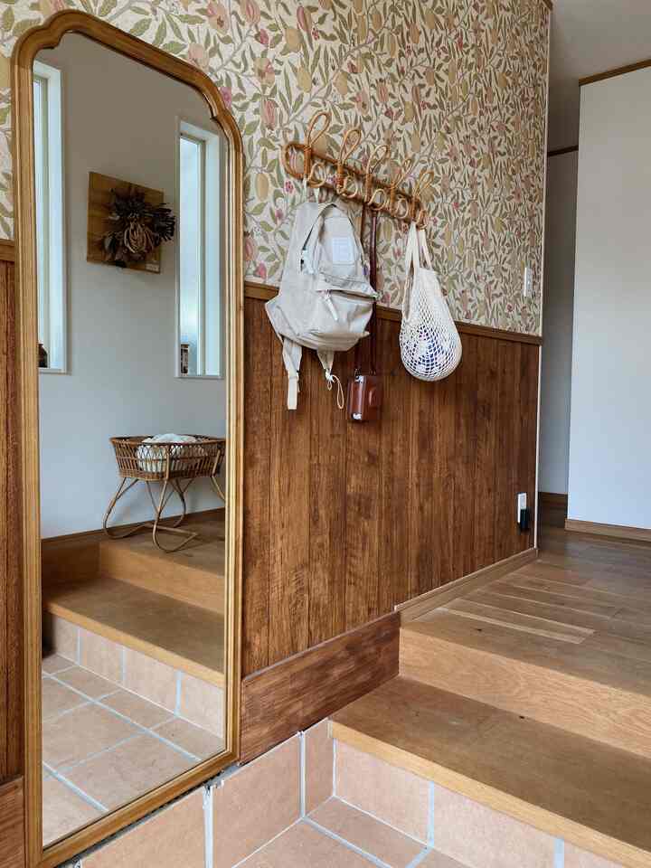 木目調の壁と花柄の壁紙があるタイル床の温かみのある玄関、鏡とコートハンガーが配置された印象的な空間