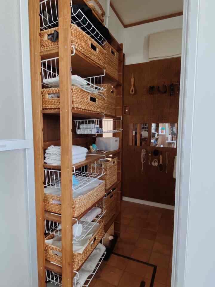 ナチュラルなブラウンの木目調が特徴的な狭い廊下に、ラタンバスケットを備えた収納ラックと壁掛けの鏡が配置された整理されたインテリア