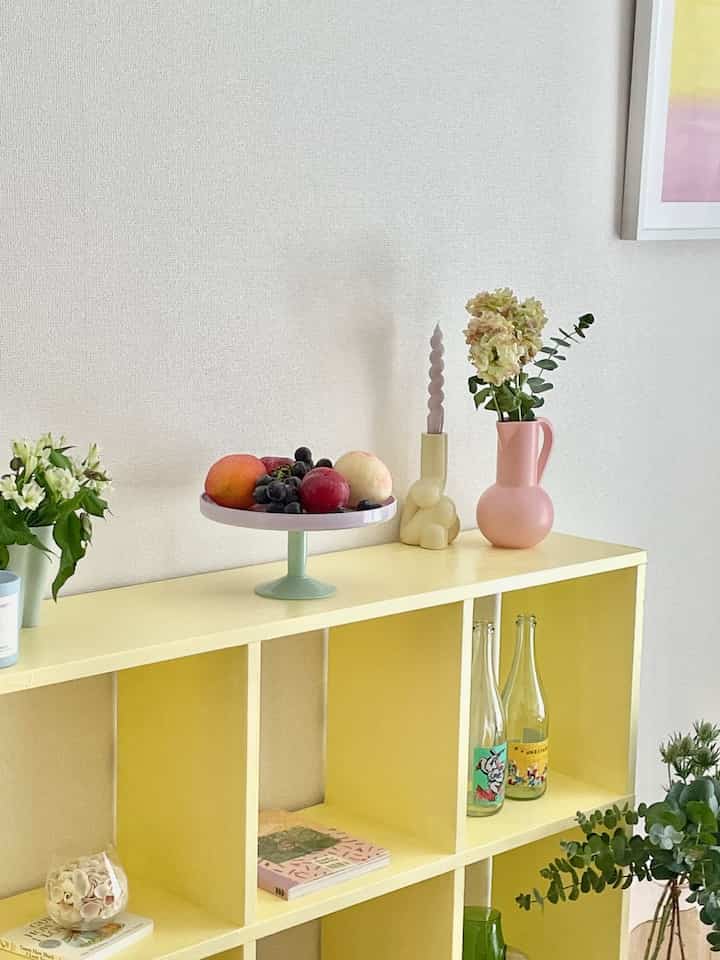 ベージュの壁と黄色の棚があるキッチン空間、花瓶とフルーツトレイが配置された明るく爽やかな印象のインテリア