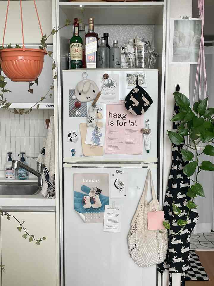 パステルカラーのアクセントがある白いキッチン、冷蔵庫にはポスターとウサギ柄のエプロン、収納用のネットバッグが掛けられたかわいい空間