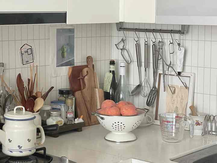 白いタイル壁と調理器具が配置されたキッチンカウンター、木製まな板と桃が入ったザルが置かれた清潔感のあるキッチンスペース