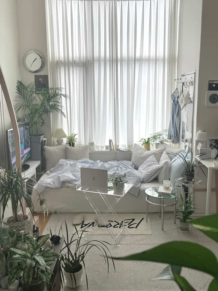 明るい白を基調としたワンルームのリビング兼寝室、中央に透明なアクリルテーブルと植物が配置されたナチュラルモダンなインテリア