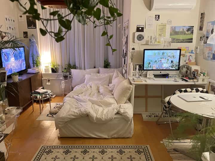 ホワイトトーンのワンルーム寝室、中央に白いベッドと右側にデスク、多数の植物とカーテンが配置された温かみのある快適な空間