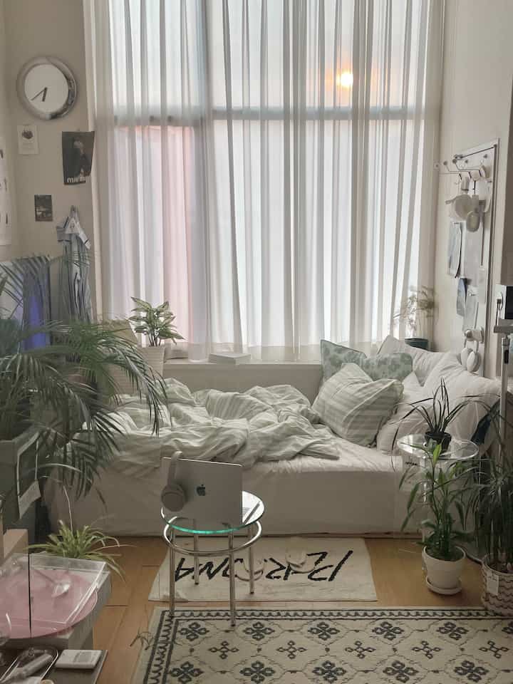 白と緑の色調の寝室、大きな窓の前にソファベッドと多くの植物が配置された落ち着いたナチュラルモダンな空間