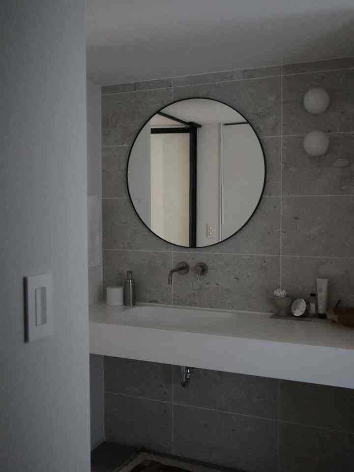 グレーのタイル壁に黒縁の丸い鏡、白い広い洗面台が配置されたシンプルで雰囲気のあるバスルーム