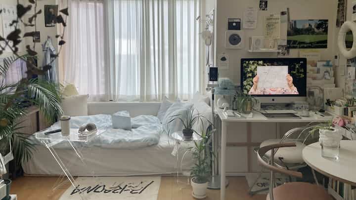 ホワイトとベージュのトーンのワンルーム、ベッドと透明アクリルテーブル、デスク、植物が配置された落ち着いたコンパクトな空間