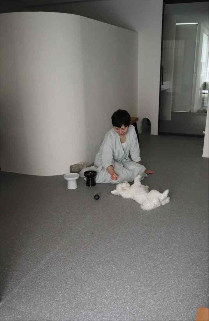 グレーの床と白い曲線壁のナチュラルな狭い部屋、床に座ってペットの犬と遊ぶ人物