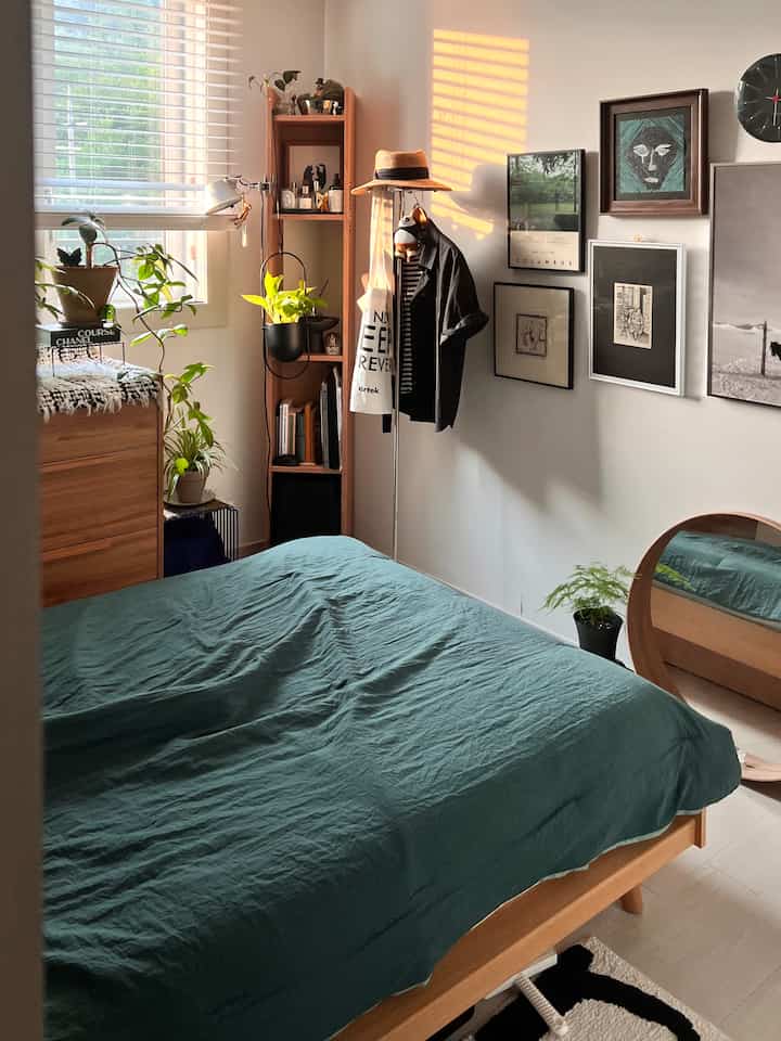 白い壁と木目調家具のある小さく整頓された寝室、緑の植物と額縁が自然な雰囲気を演出している