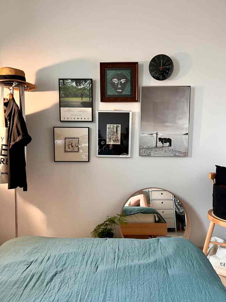 白い壁と木目調の自然な要素が調和した寝室、壁に額縁と時計が配置され、ティール色のベッドカバーでシンプルで落ち着いた雰囲気のインテリア