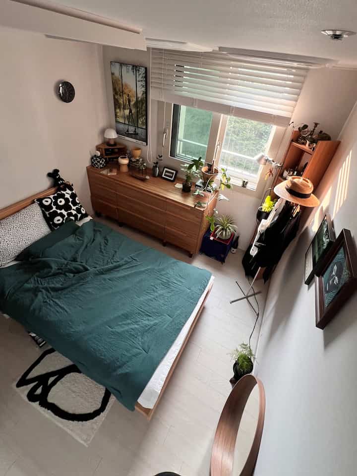 白い壁と木目調の家具が配置されたナチュラルな寝室、緑の寝具と複数の植物が調和した心地よい空間