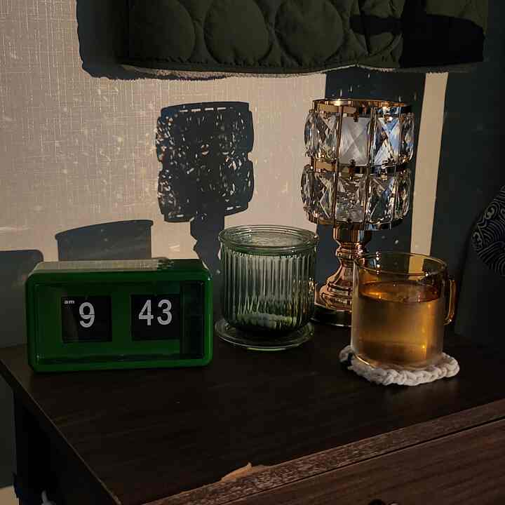 落ち着いたダークトーンとブラウンの木製ナイトテーブルの上に、緑色のフリップクロック、透明の花瓶とカップ、クリスタルランプが印象的に配置された雰囲気のあるインテリア