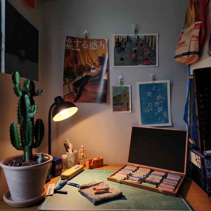 暖かい照明の白い壁と机があるアートワークスペース、サボテンの鉢植えと壁にかかった水彩画が配置された雰囲気のあるインテリア