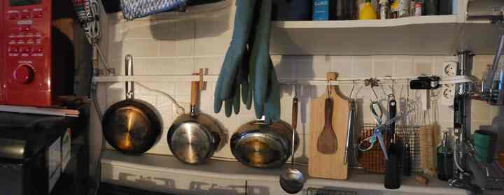 ナチュラルな色調のキッチン、ガス管と食器乾燥棚の柱にストラップでフライパンや鍋、調理器具を収納した実用的で生活感のあるインテリア