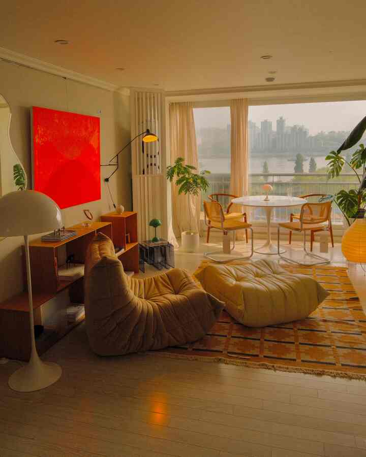 ベージュと赤の色調のリビング、ミッドセンチュリーモダンの家具と大きな窓から川辺の景色が見える暖かい雰囲気のあるインテリア