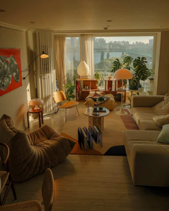 暖かいウッドトーンとベージュのリビング、大きな窓から自然光が入り、ソファとスツールが配置された居心地の良い空間