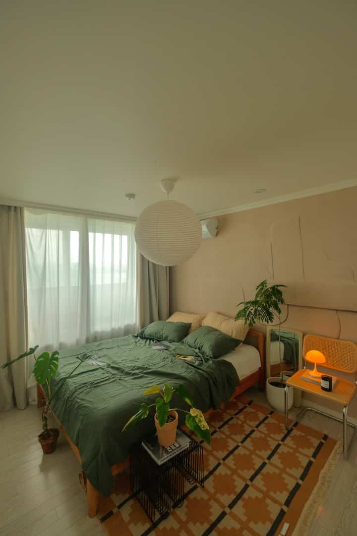 ベージュの壁とグリーンの寝具が印象的なナチュラルスタイルの寝室、木製ベッドと植物が配置された落ち着いたインテリア