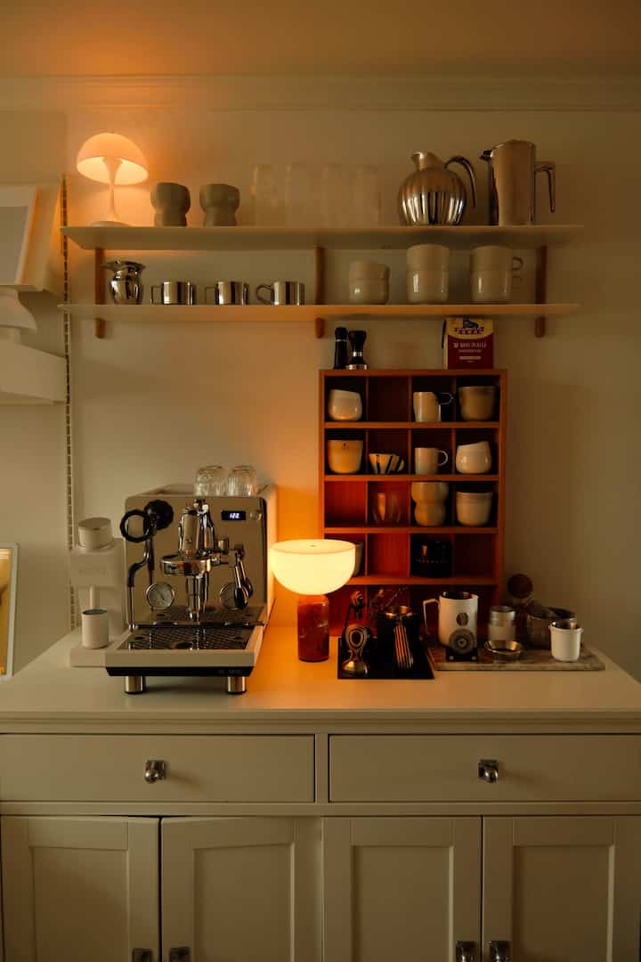 温かみのあるウッドトーンと白を基調としたミニマルなおうちカフェ空間、テーブルランプとエスプレッソマシンが配置された落ち着いたセルフケアコーナー
