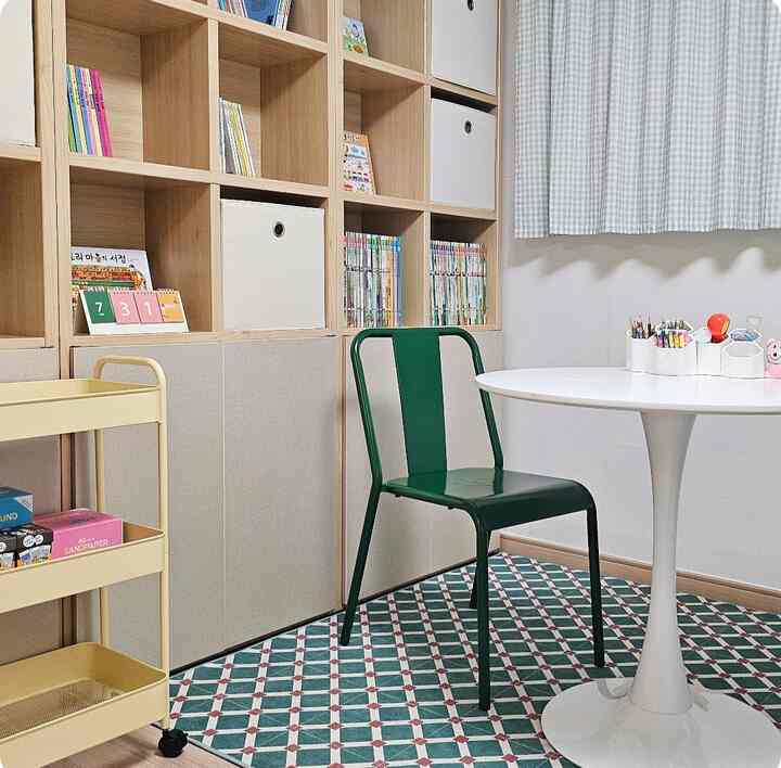 白とナチュラルウッドトーンの小さなスタディルーム、本棚と白い円形テーブル、緑の椅子が配置された落ち着いた空間