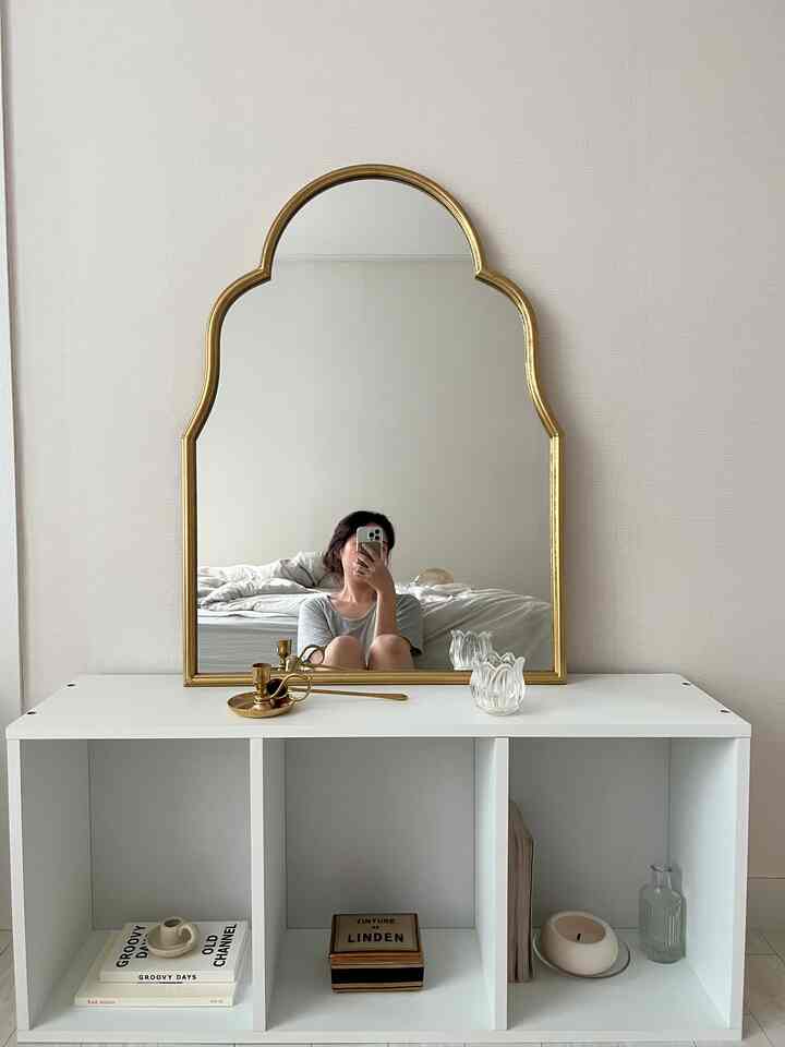 ホワイトとゴールドの色調のリビングルーム、ゴールドフレームの鏡とホワイトの本棚にキャンドルが配置された落ち着いたコテージスタイルのインテリア