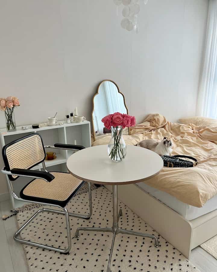 白い壁とベージュの寝具がある寝室、ラタンの椅子と花瓶が置かれた小さな丸テーブルが配置されたシンプルでナチュラルな雰囲気のインテリア