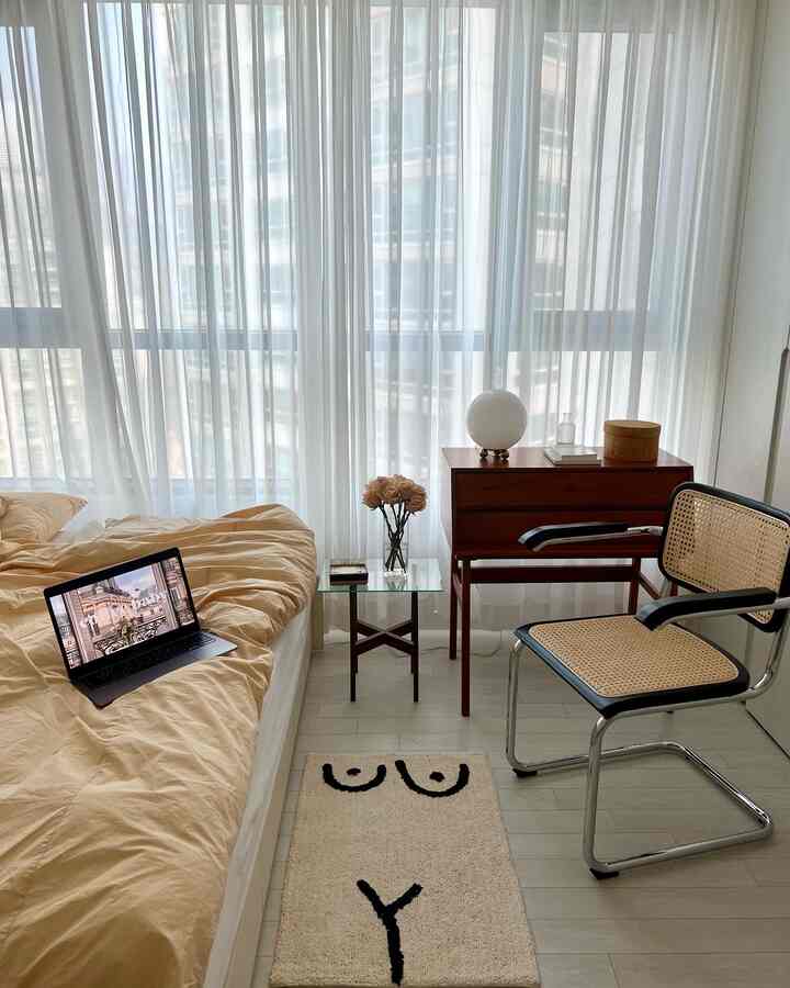 ベージュトーンの寝室、中央左にベッドとノートパソコン、右側に木製の引き出しとラタンチェアが配置されたシンプルな自然な雰囲気のインテリア