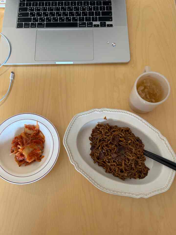 ナチュラルな木目のダイニングテーブルに黒い짜파게티、キムチの白い器、ベージュのマグカップが配置された雰囲気のあるホームダイニング