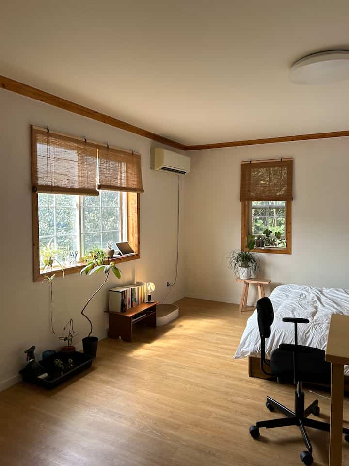 白い壁とナチュラルな木調の6畳寝室、白いベッドと黒いオフィスチェア、植物が配置された落ち着いた印象の空間