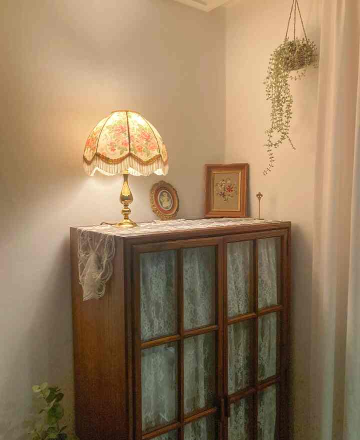 柔らかなベージュの壁と木製キャビネットが配置された空間、花柄のテーブルランプとレースカーテンが居心地の良い雰囲気を演出