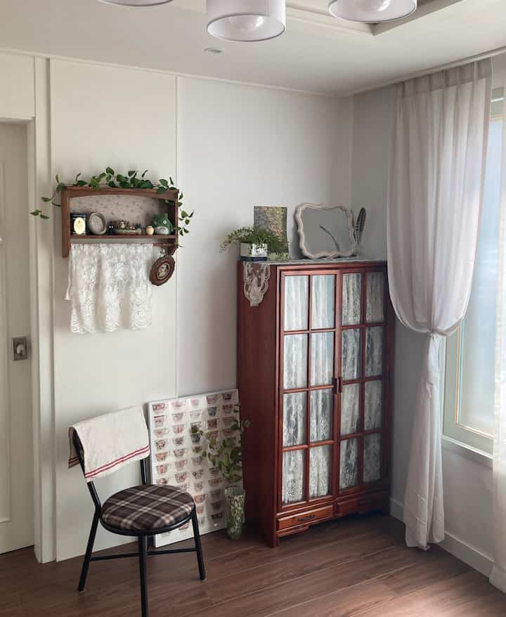 白い壁と茶色の木製床が調和するリビング、収納キャビネットと壁掛け棚、植物が自然に配置された雰囲気のある空間