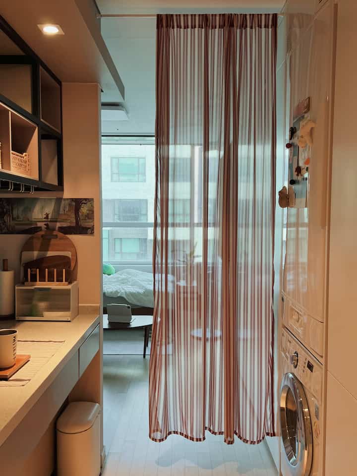 ホワイトトーンの狭く縦長のキッチンと洗濯スペースが玄関通路とつながり、中央に透けたストライプのカーテンが配置されたインテリア