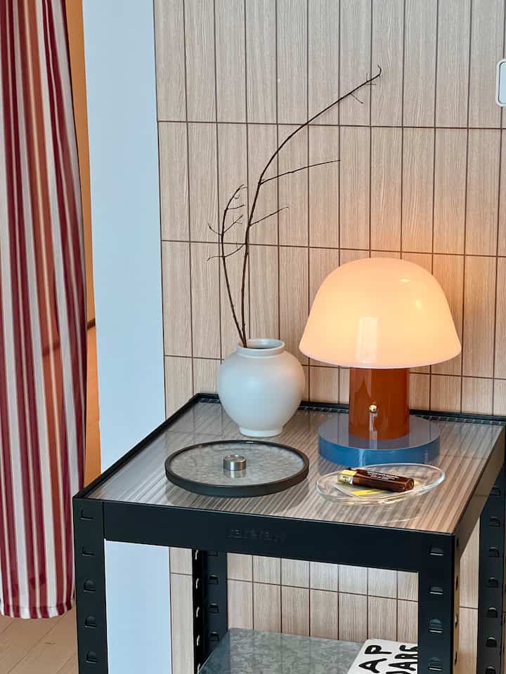 ナチュラルトーンの壁と黒いメタルのサイドテーブルの上に白いマットな花瓶とオレンジのテーブルランプが置かれた温かみのある作業スペース