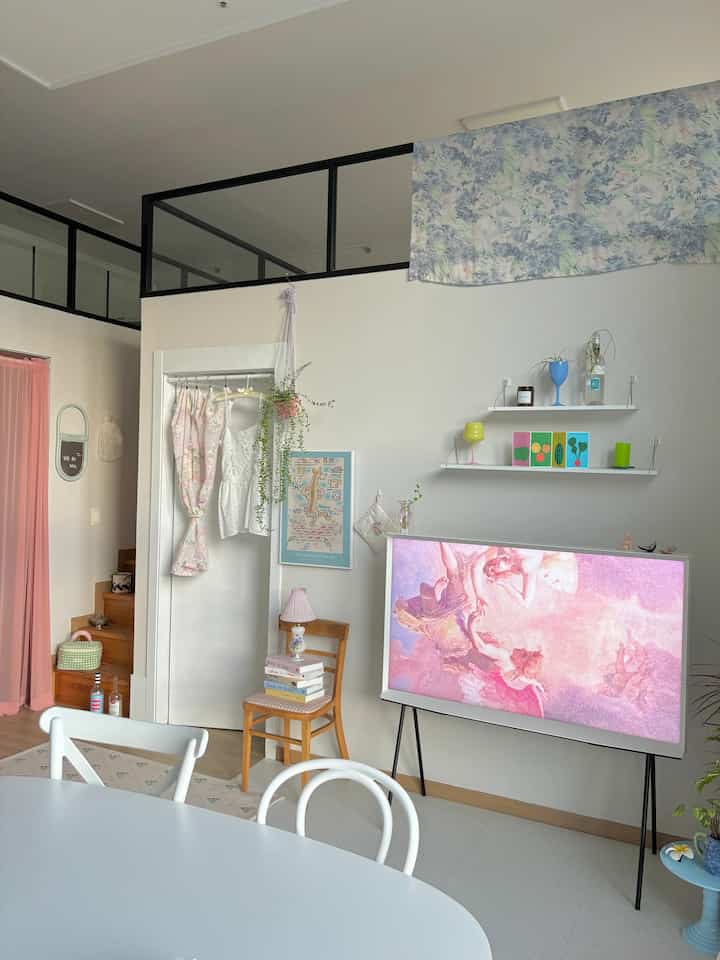 ホワイトとピンクの色調のリビング、テレビスタンドとピンクのテーブルランプ、壁の棚に飾りが配置された雰囲気のある空間
