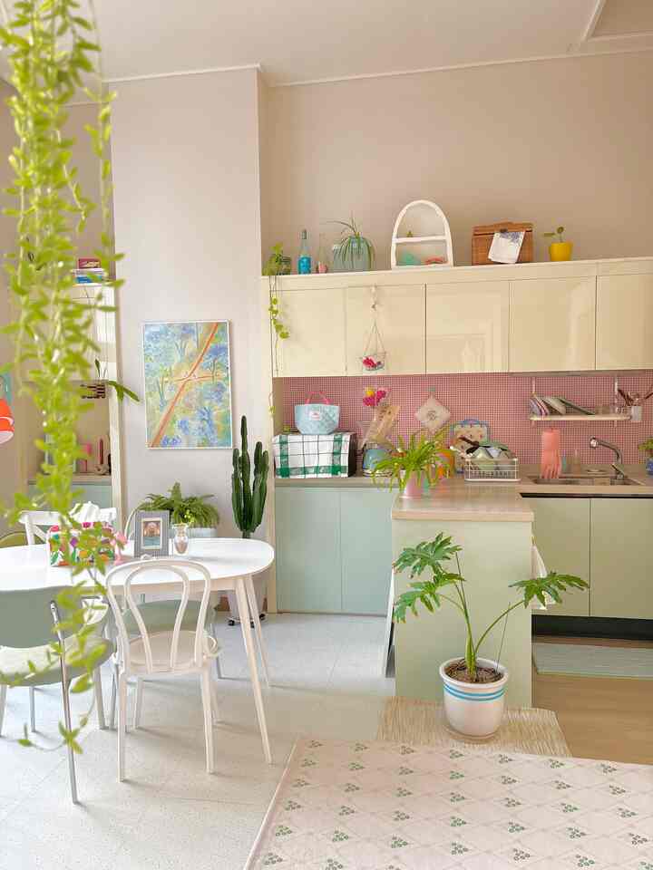 ピンクと赤のタイルが映えるミント色のキャビネットがある明るいキッチン、白いダイニングテーブルと植物が配置された空間