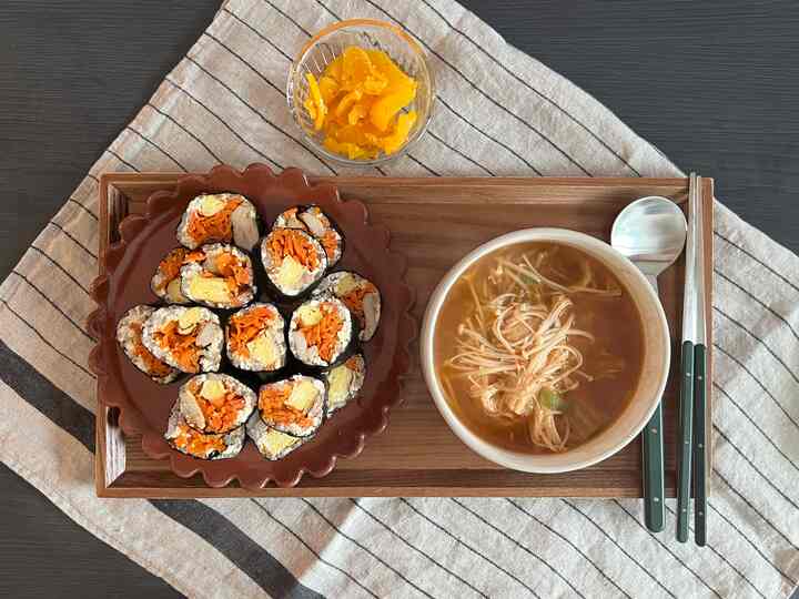 ナチュラルカラーのテーブルに木製トレイが置かれ、キンパの皿、スープの器とカトラリーが整然と配置された食事セット