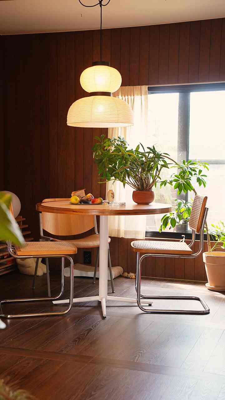 濃いブラウンの木目壁と木製丸テーブルと椅子、ペンダントライトと白いカーテンが調和した温かみのあるダイニング空間