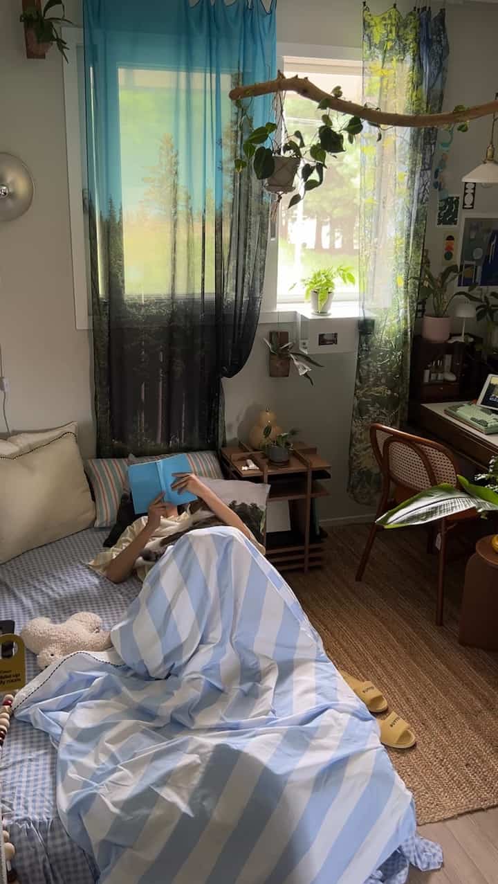 青と自然柄のカーテンと植物がある居心地の良い寝室で、一人の人が猫と一緒にベッドで本を読んでいる雰囲気のあるインテリア