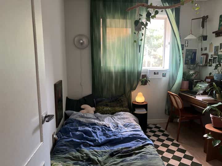 緑のカーテンと木目調家具が配置された、日差しの入る居心地の良い一人用寝室