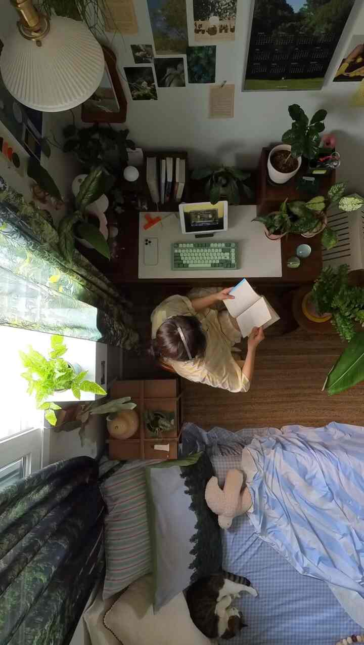 ナチュラルなグリーントーンの寝室、デスクと多様な植物が配置され、猫と人が本を読む雰囲気のある空間