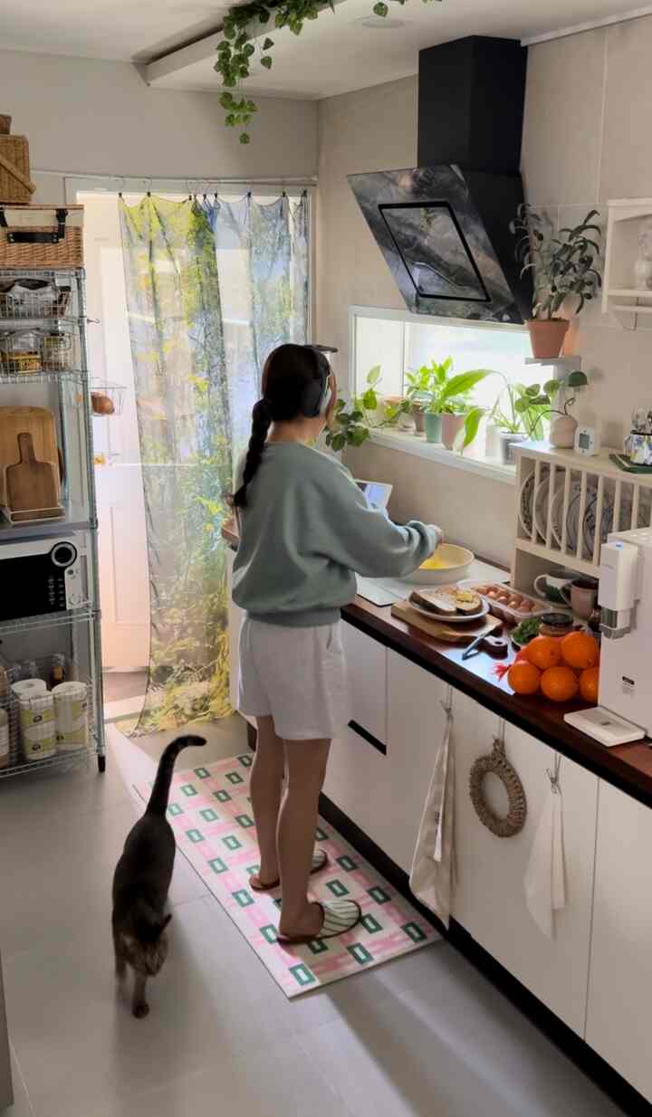 ホワイトの下部キャビネットと木目調カウンターが特徴的な縦長のキッチン、窓際の植物と猫がいる雰囲気のあるインテリア