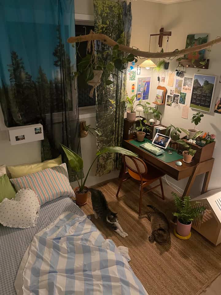 緑の植物が豊富な木目調のワンルーム寝室、机とベッド、2匹の猫がいる居心地の良い空間