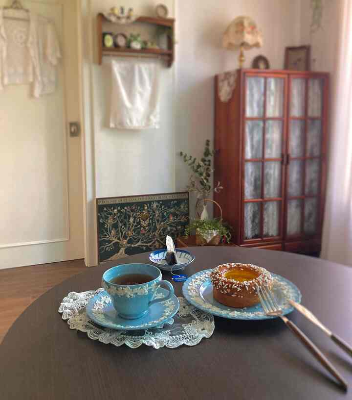 ナチュラルな色調のダイニング、木製丸テーブルに青いウェッジウッドのティーカップと皿、レースのテーブルランナーが配置された雰囲気のある空間