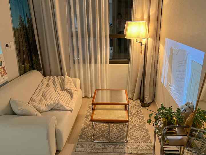 ベージュとナチュラルカラーの小さいリビング、クリーム色のソファと柄のラグ、プロジェクターとフロアランプが配置された雰囲気のあるインテリア