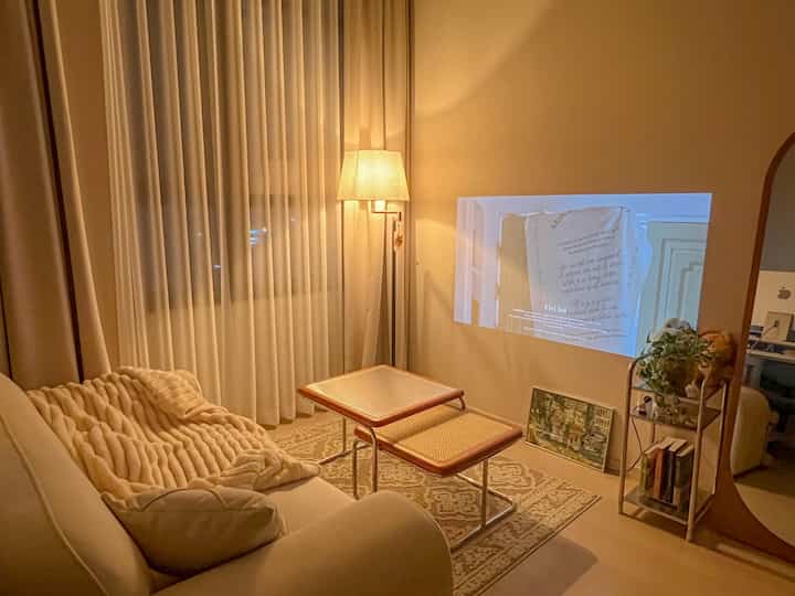 暖かいベージュトーンのリビング、ベージュのソファと木製サイドテーブル、フロアランプ、壁に映し出されたプロジェクターが落ち着いた雰囲気を作る空間