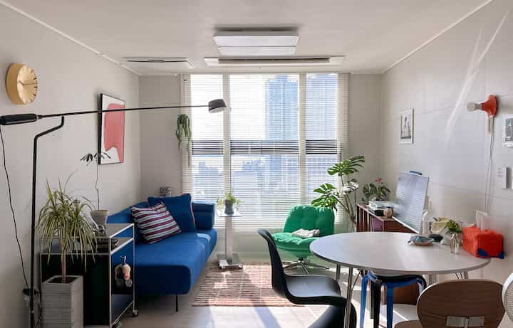 白と青の色調のリビング、青いソファと緑のアームチェア、大きな窓のブラインドが配置された明るく居心地の良い空間