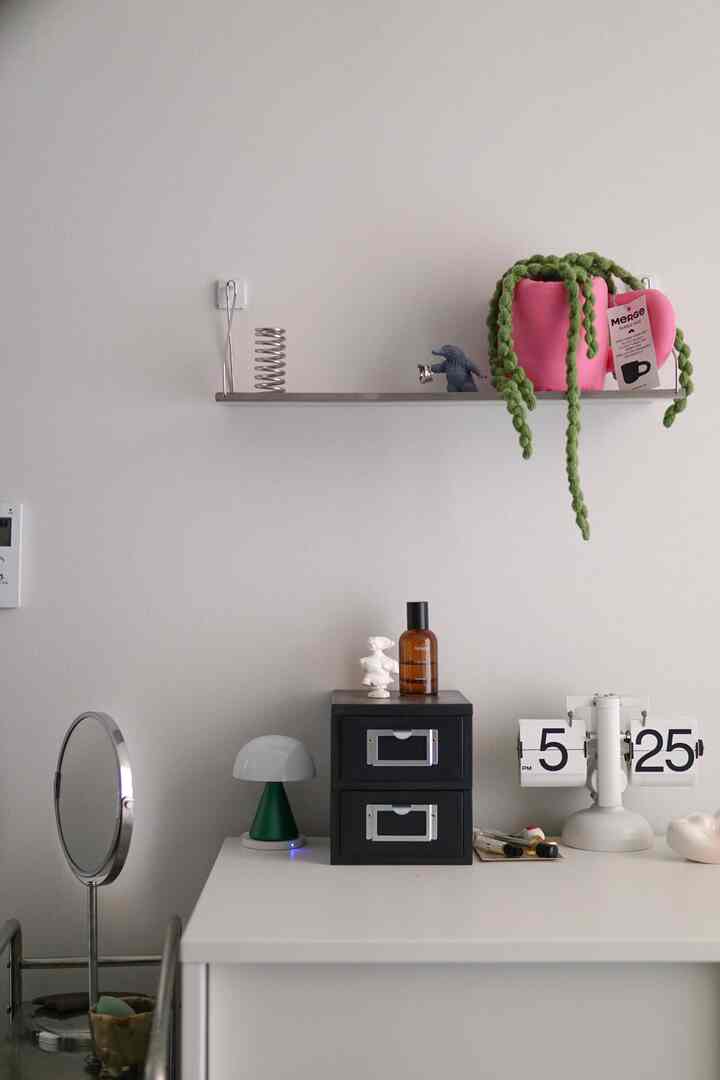 ホワイトの壁とデスクの上にブラックのミニ引き出し、メタル棚にピンクのファブリックプラントが置かれたモダンな寝室空間