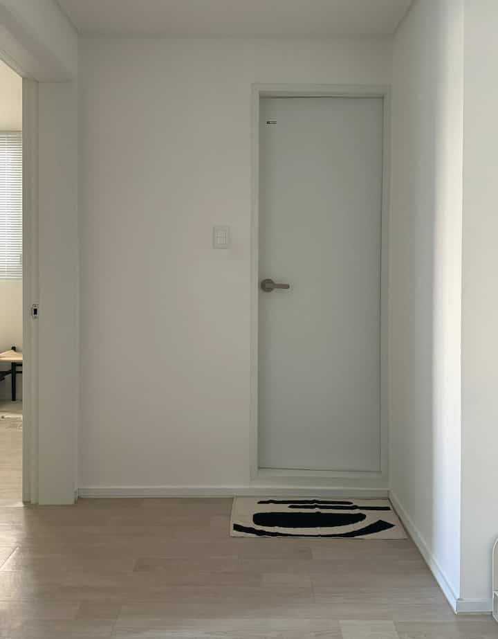 ホワイトとウッドトーンの調和が取れた玄関、清潔感のあるトイレドアとシンプルな模様のラグが配置されたモダンな雰囲気