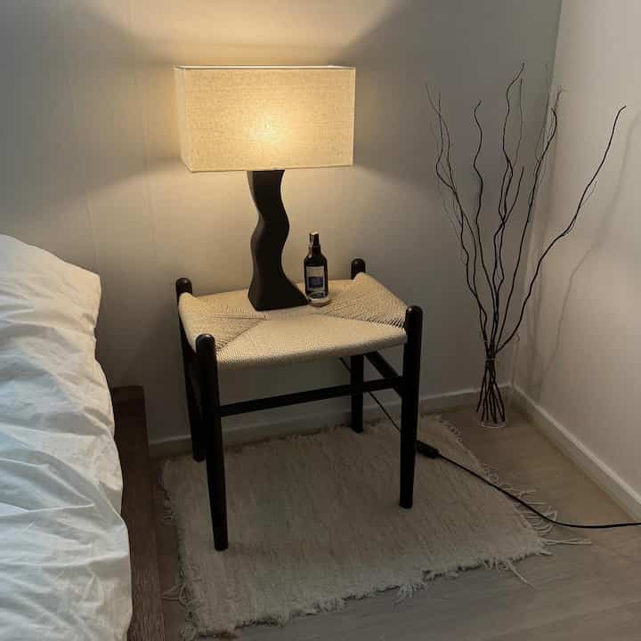 ベージュとライトブラウンのトーンの寝室の一角にスツールと照明が置かれた、自然な雰囲気のあるモダンなインテリア