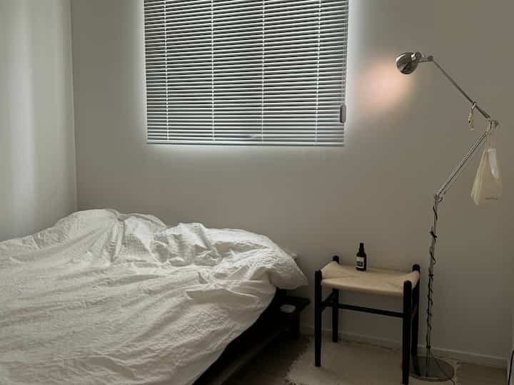 白とベージュの色調の寝室、ベッドとスツール、フロアランプが配置されたシンプルで落ち着いた印象的な空間